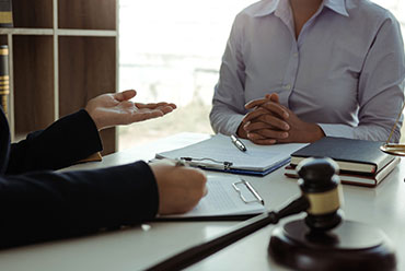 A man sits at a desk with a lawyer discussing his case, with a gavel, legal book, and clipboard with papers on the table.
